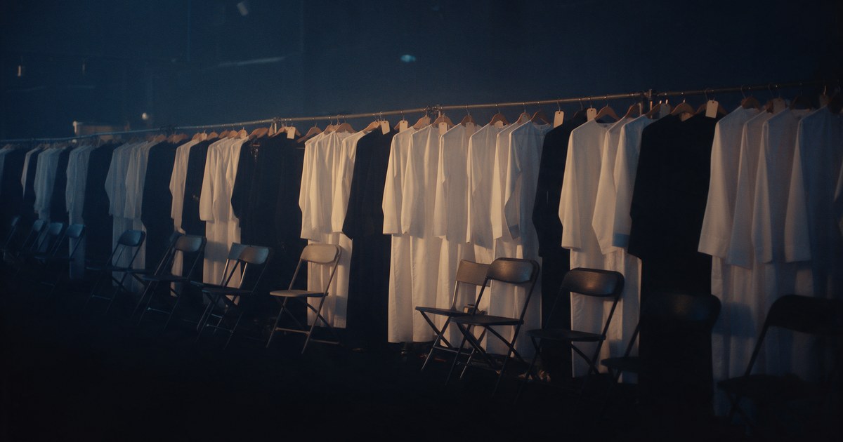 A wardrobe rail of white thobes and black abayas beside rows of empty folding chairs in a holding area