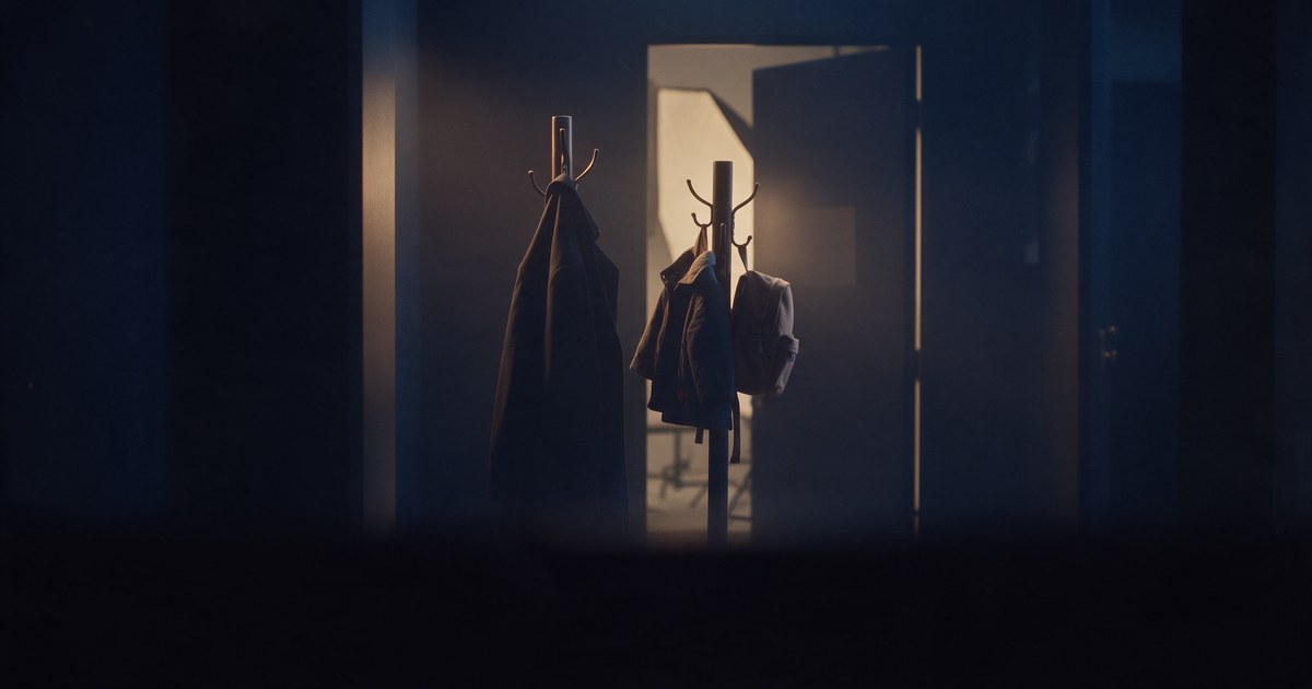 An adult coat beside a child-size jacket and backpack on coat stands at an open photo studio door